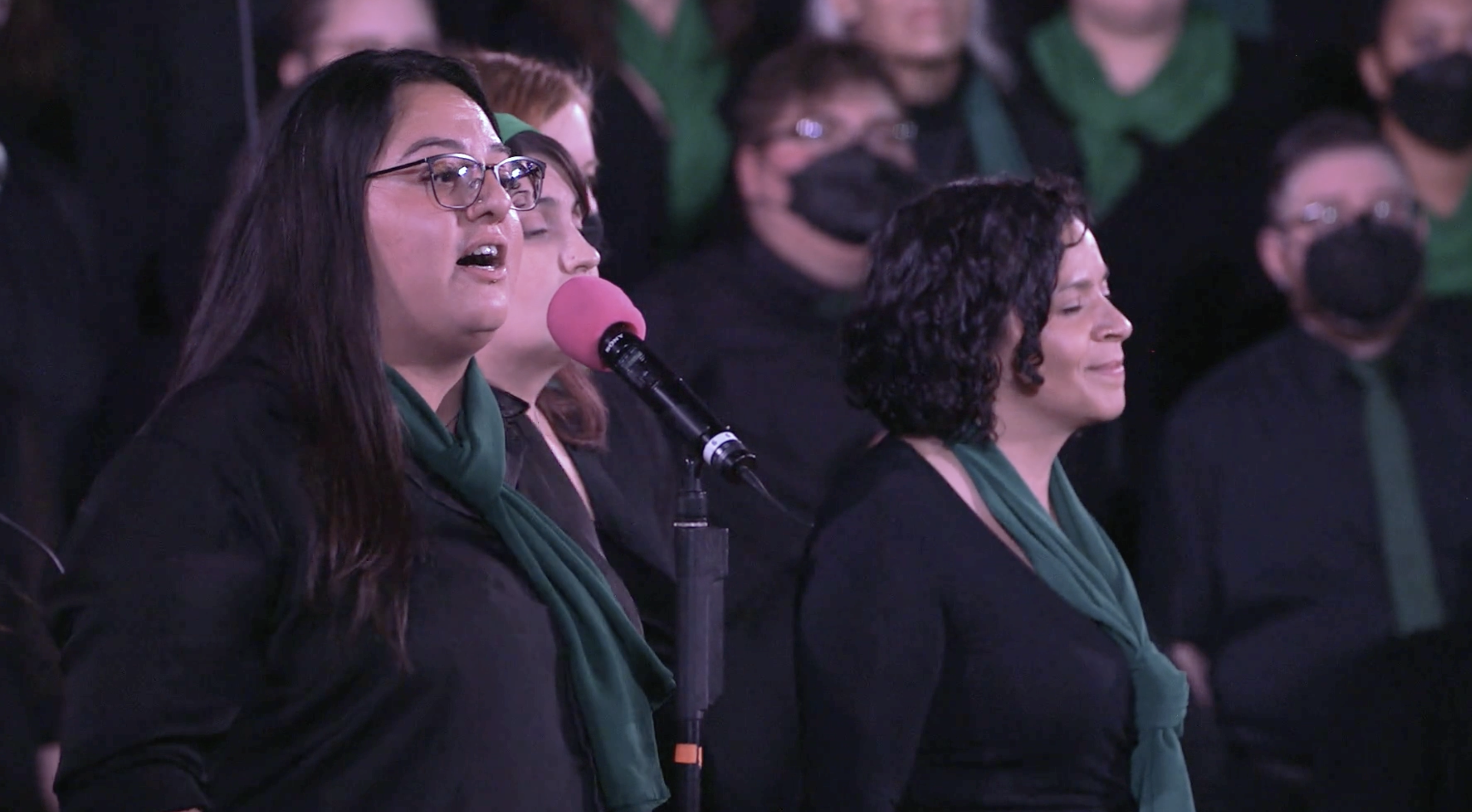 A soloist with long, dark hair sings into a microphone behind a choir, all wear black with green scarves.